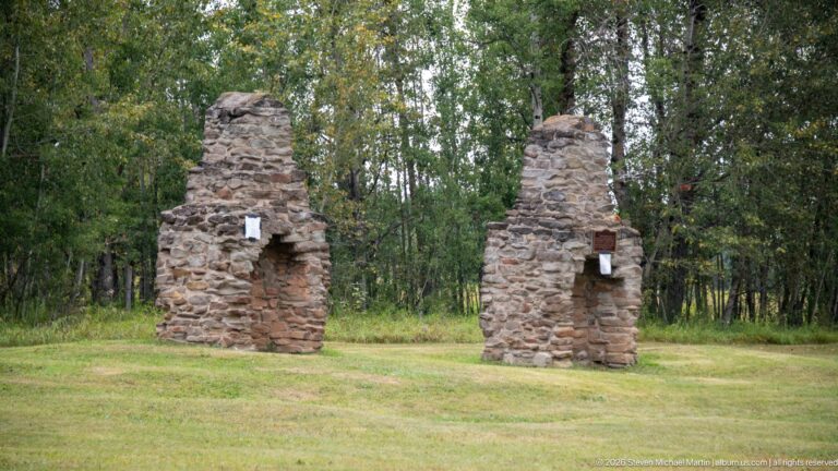 Two freestanding rough-stone chimneys with hearth openings standing on grass in front of a wooded backdrop.