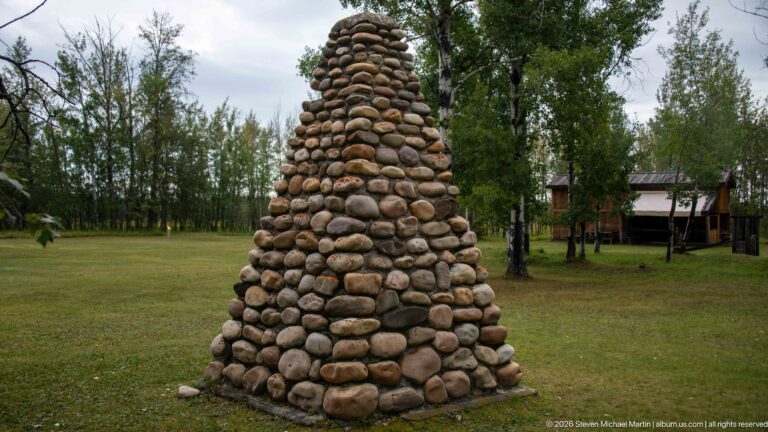 Tall tapered monument of rounded stones standing on a grassy lawn with trees and buildings behind.