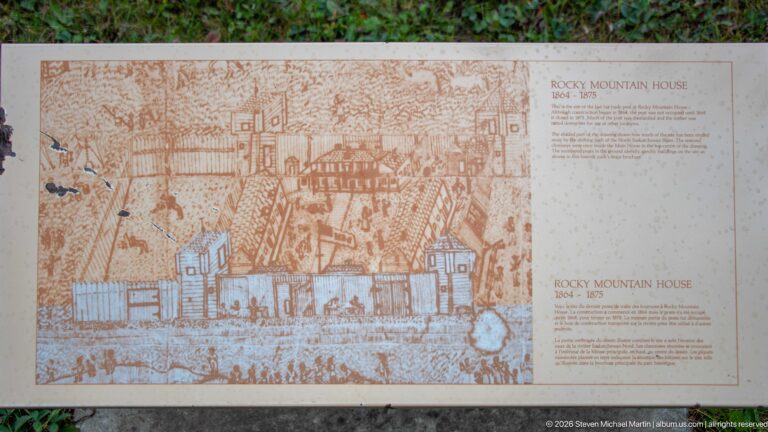 Weathered interpretive panel with sepia historical drawings of fenced timber buildings and figures at Rocky Mountain House.