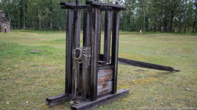 Freestanding dark wooden apparatus with tall vertical posts, crossbars, rope, and a lower sliding or weighted component.