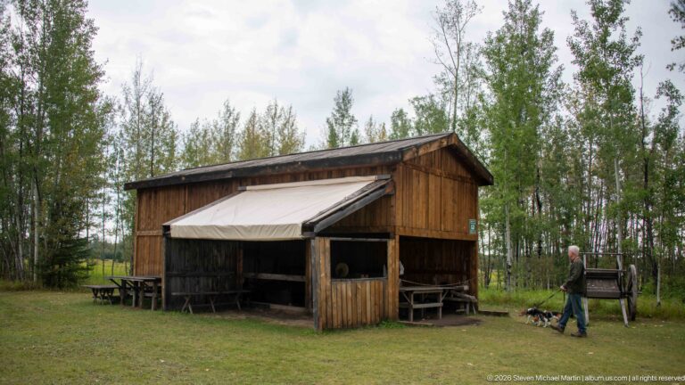 Small timber workshop with open front, pale canvas awning, benches, and a visitor walking across the grass.