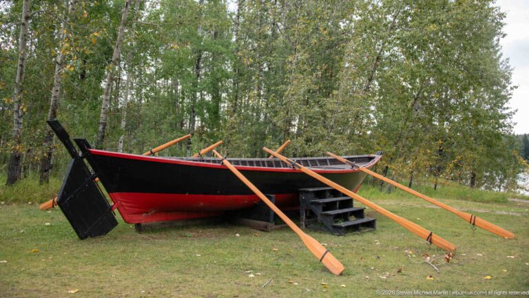 Large black-and-red wooden boat on grass with several long wooden oars extending across the hull.