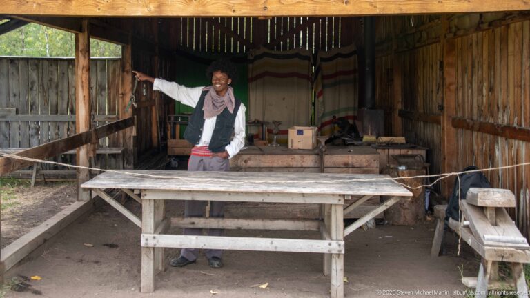 Costumed interpreter gesturing behind a long wooden table inside a rustic workshop filled with boxes, barrels, tools, and blankets.