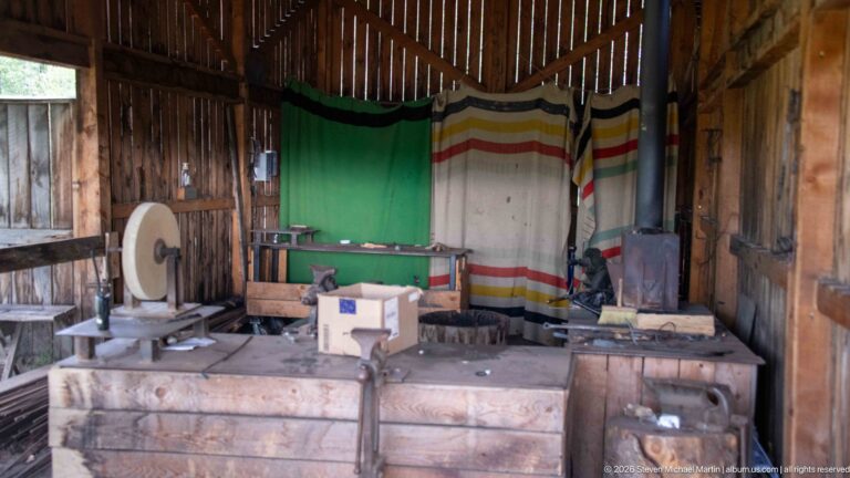 Rustic wooden workshop with grindstone, workbench, iron stove, boxes, tools, and striped blankets hanging across the rear wall.