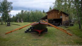 Large dark wooden boat on supports with long orange-brown oars, positioned in front of a timber building and wheeled equipment.
