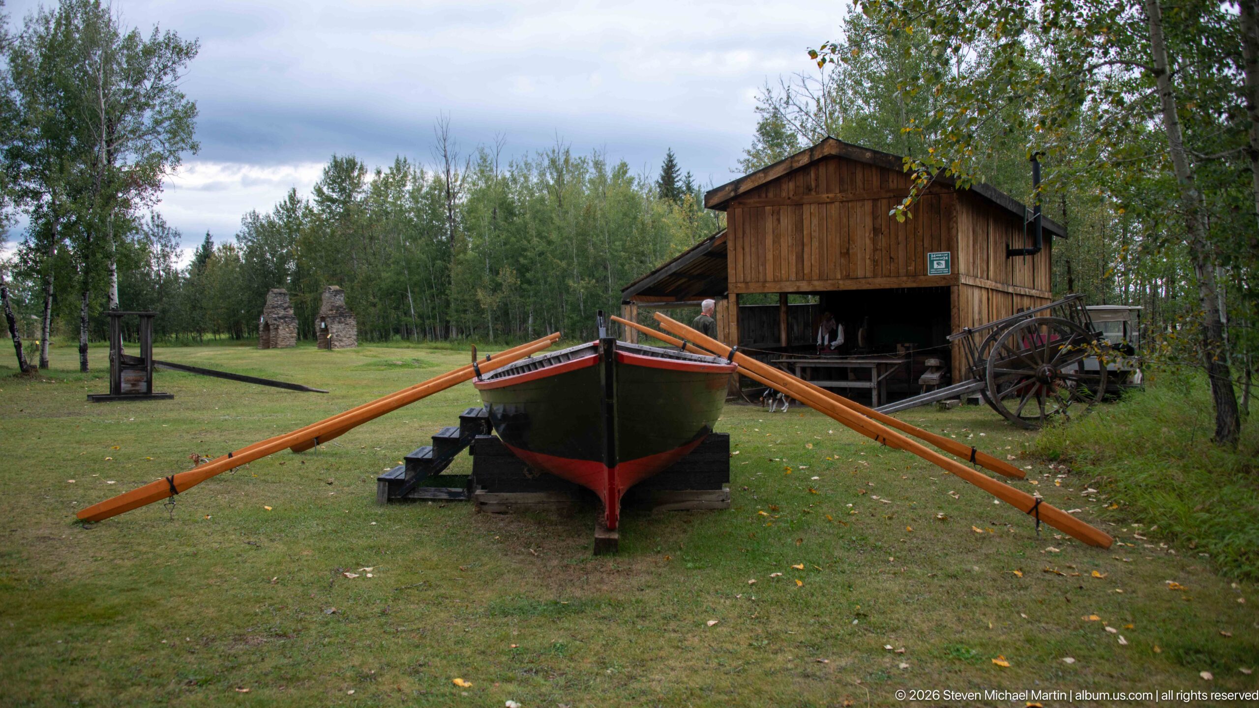 Large dark wooden boat on supports with long orange-brown oars, positioned in front of a timber building and wheeled equipment.