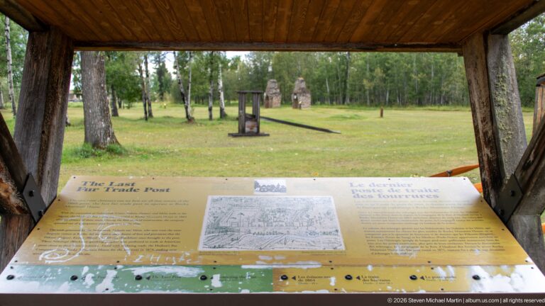 Timber-roofed outdoor interpretive station with a sloping panel, open lawn, trees, and distant stone features beyond.
