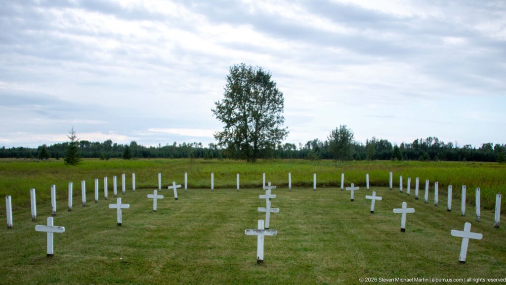 Wide grassy burial ground with numerous white cross-shaped and upright grave markers, bordered by meadow and trees.