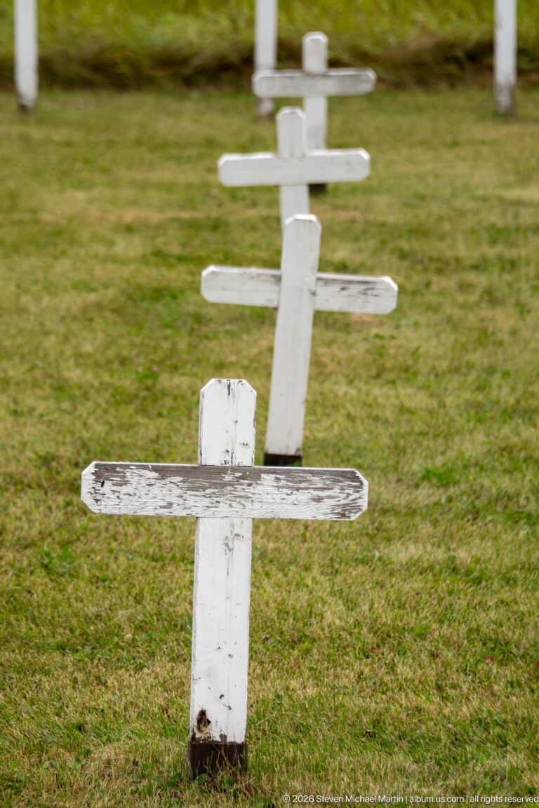Weathered white wooden cross in the foreground with additional cross-shaped grave markers receding across grass.