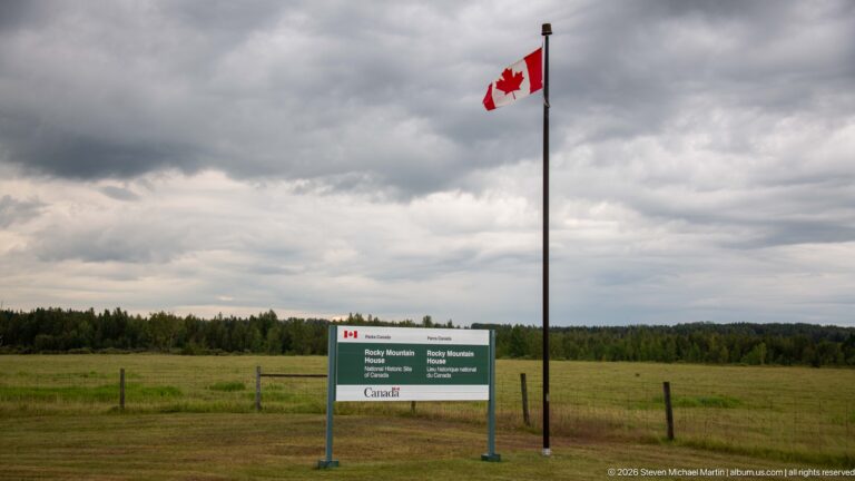 Canadian flag on tall pole above bilingual Rocky Mountain House National Historic Site sign in open grassland.