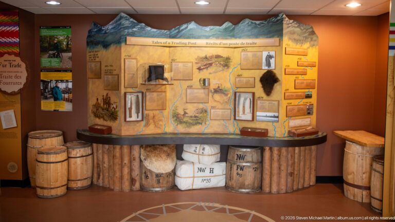 'Tales of a Trading Post' exhibit with illustrated map, wooden barrels, wrapped bales, small artifacts, and interpretive panels.
