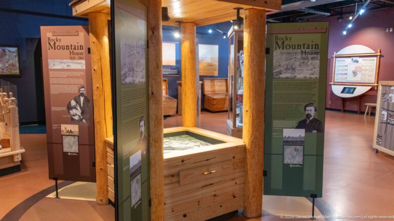 Visitor-centre gallery with timber posts, tall interpretive panels, portraits, maps, and a low wooden exhibit case.