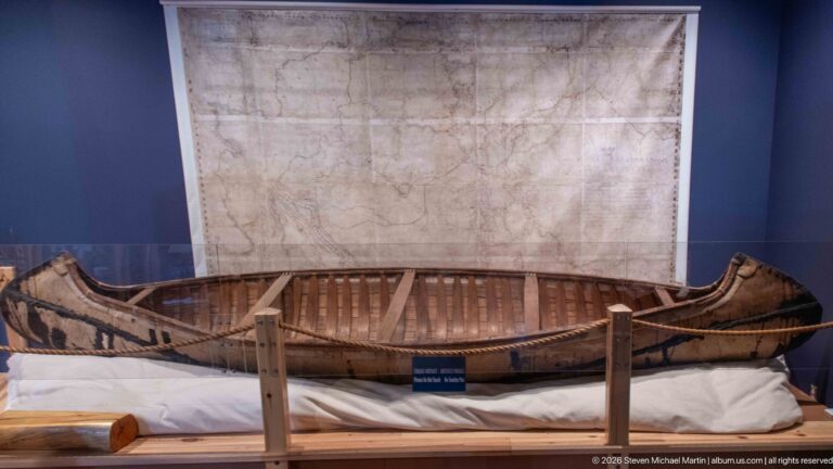 Long bark-covered Indigenous watercraft with exposed ribs and raised ends displayed beneath a large historic map.