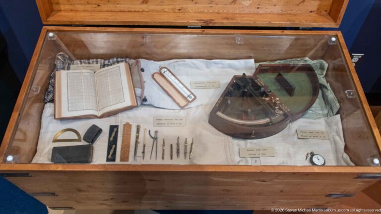 Glass display case with an open ledger, measuring tools, writing implements, navigation instrument, and other small historical objects.