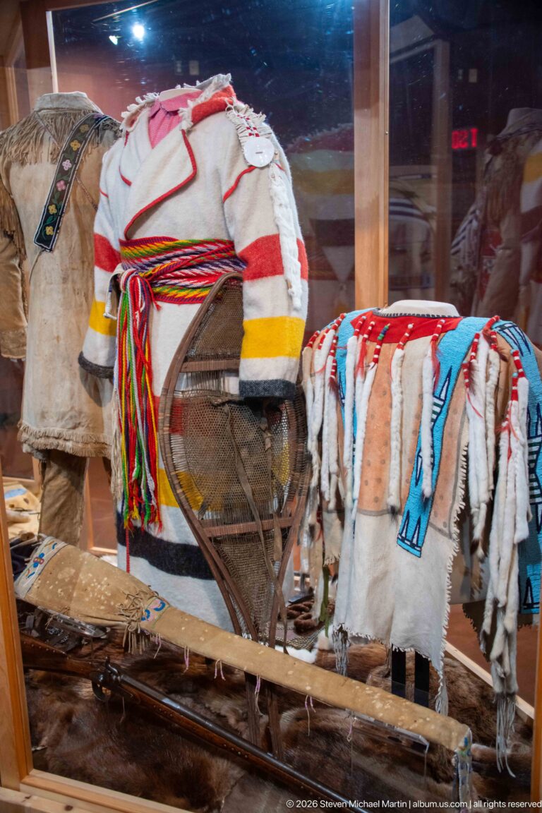 Curved wooden transport frame beside fringed and decorated hide garments in a museum display case.