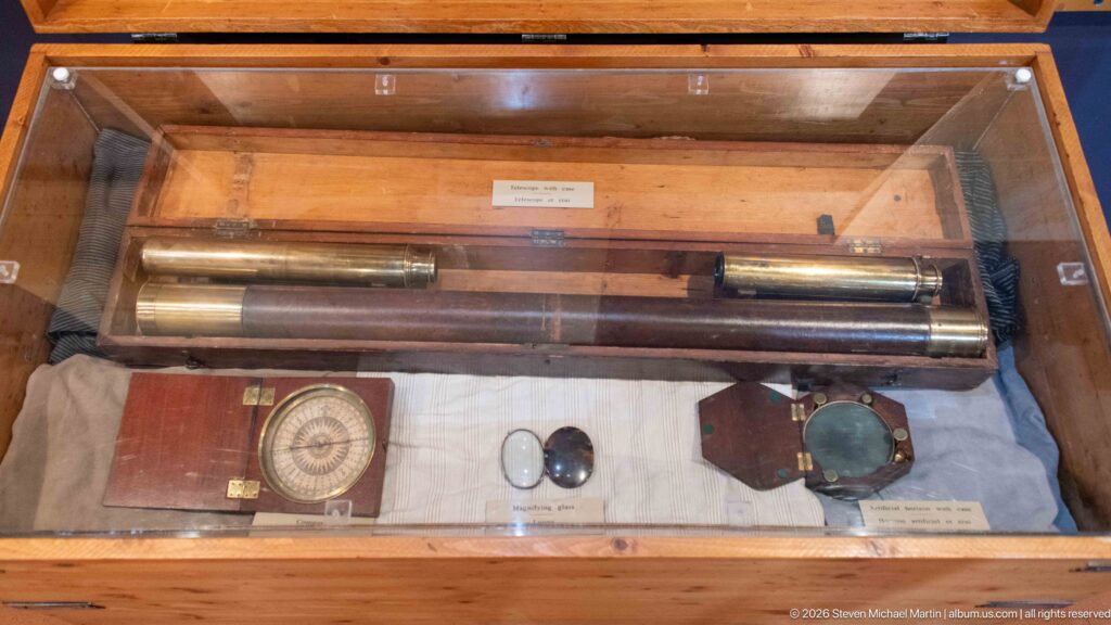 Brass cylindrical instruments, a boxed circular compass-like device, and smaller measuring tools displayed in a glass case.