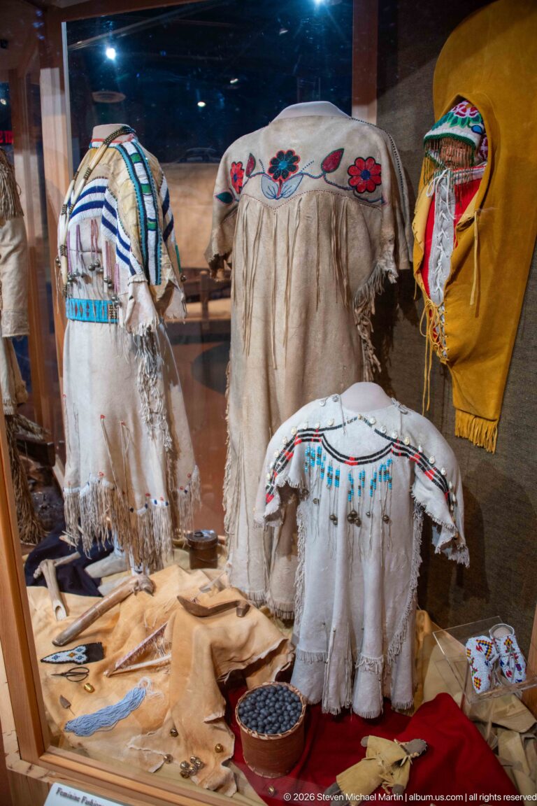 Several pale hide garments with beadwork, colorful motifs, and long fringe displayed together behind glass.