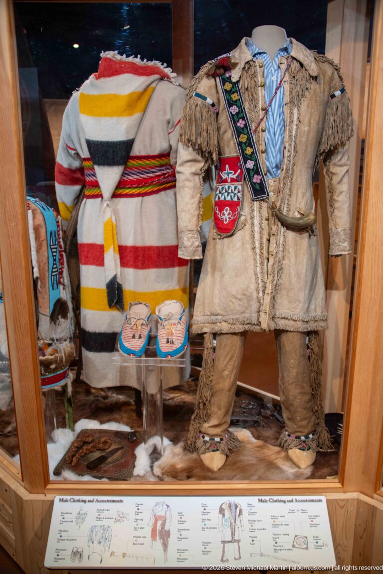 Two decorated Indigenous outfits with fringe, beadwork, moccasins, and accessories displayed behind glass.