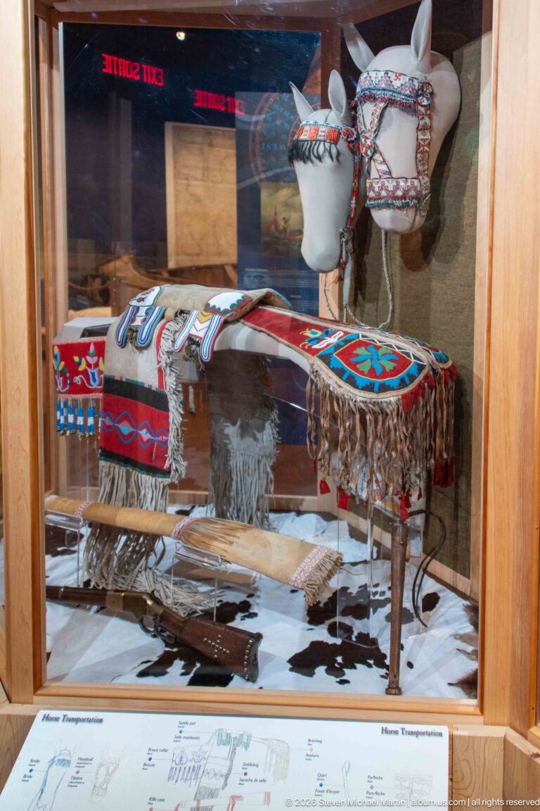 Beaded and fringed horse equipment with geometric designs displayed on a white horse-form mount behind glass.