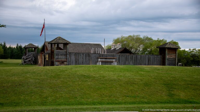 Outer side of a timber palisade with roofed corner structures on a grassy rise under clouds.