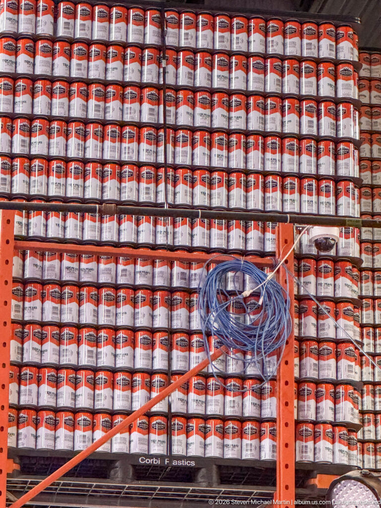 Rows of labeled beer cans fill orange shelving behind a coil of blue tubing.