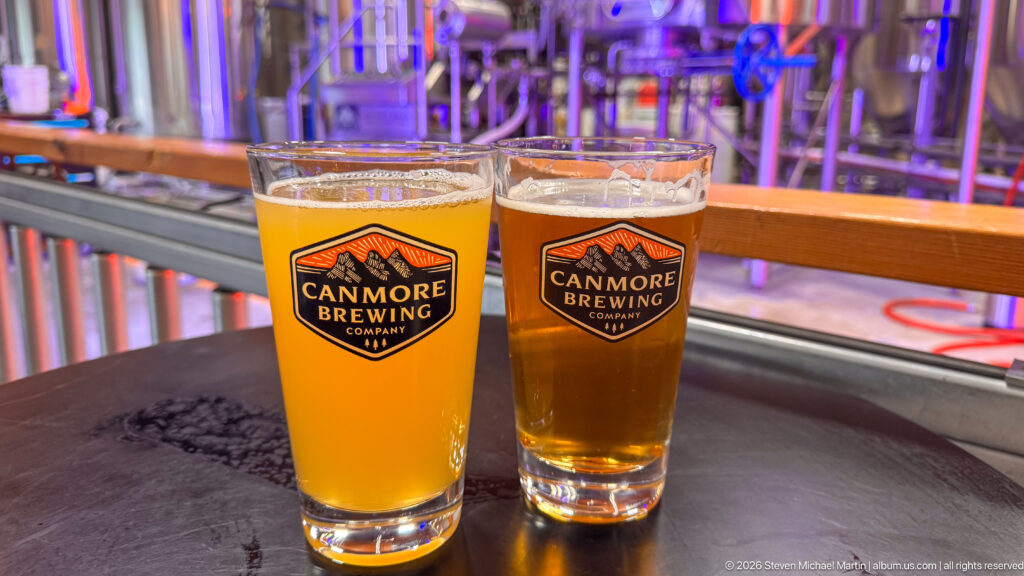 Two differently colored beers in Canmore Brewing Company glasses stand before brewery equipment.