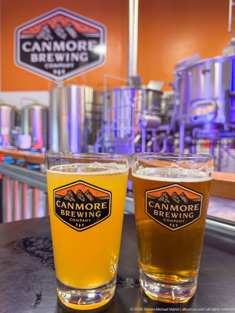 Two Canmore Brewing Company glasses hold a pale hazy beer and a darker amber pour.