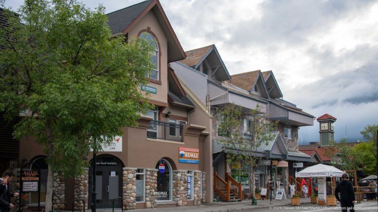A clock tower rises above gabled shops and visitors along a tree-lined street.