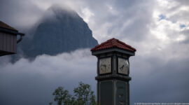 A green clock tower stands before a steep mountain slope veiled in cloud.