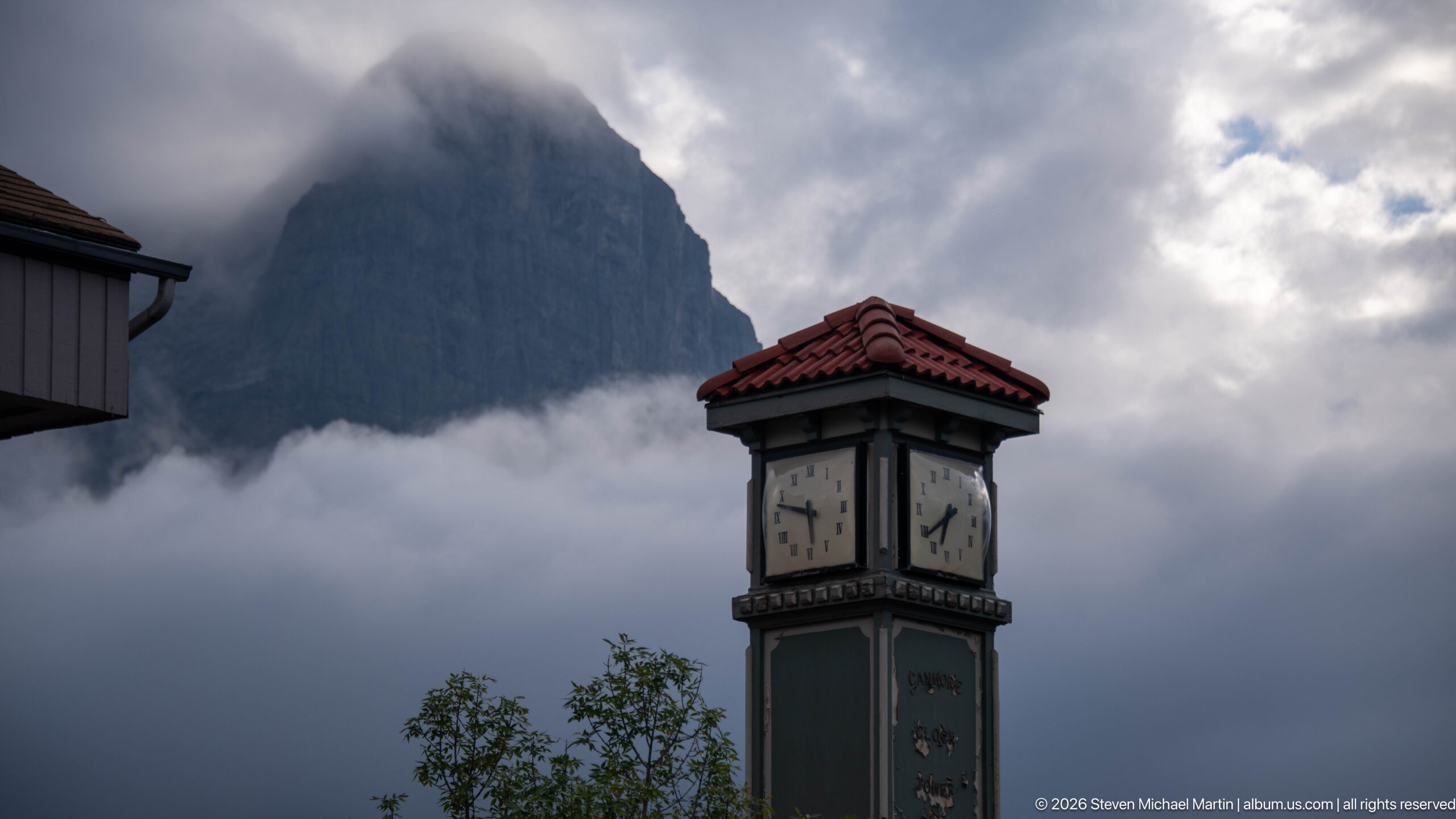 A green clock tower stands before a steep mountain slope veiled in cloud.