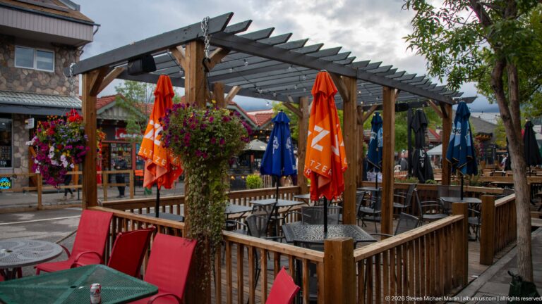 A timber pergola shelters patio tables, colorful umbrellas, and hanging flower baskets.