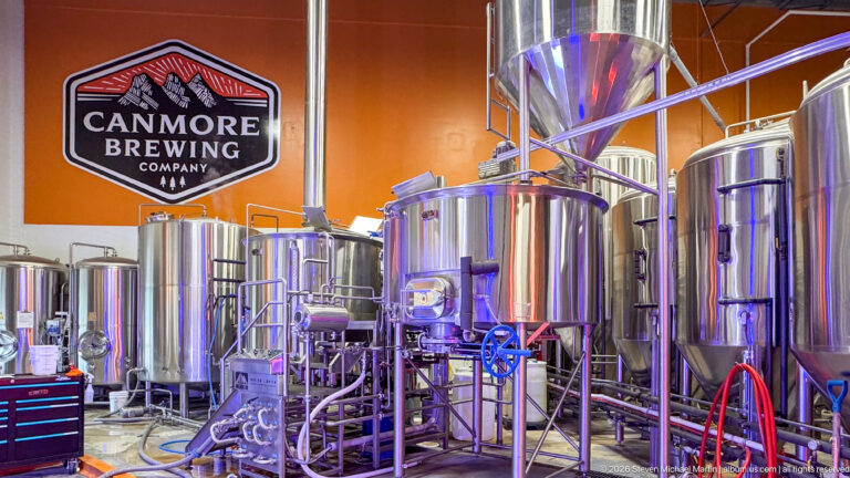 Canmore Brewing Company’s wall sign hangs above stainless tanks, pipes, and colored hoses in the brewhouse.