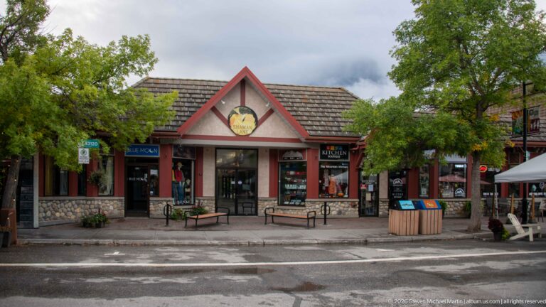 A red-trimmed, stone-fronted shop sits beneath a tiled gable on a tree-lined street.