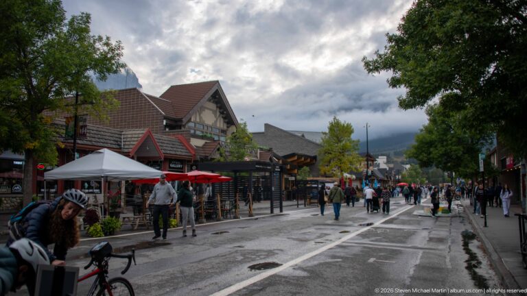 Visitors walk among timber-fronted shops and patio umbrellas along a Canmore pedestrian street.