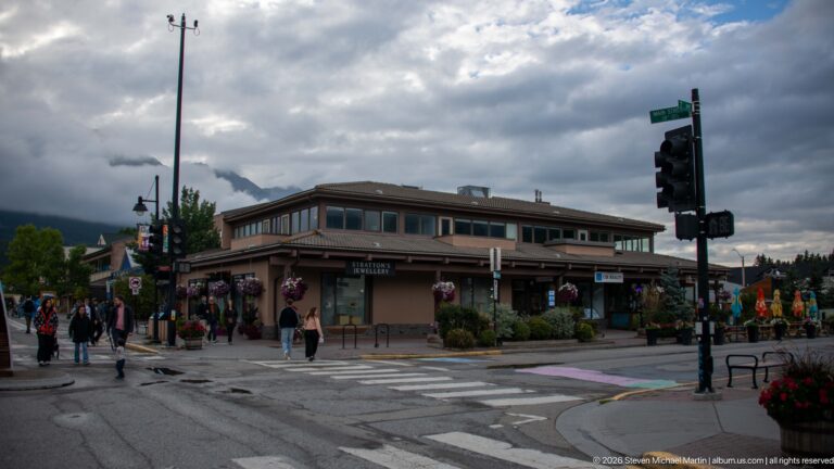 Pedestrians cross a wet intersection beside a broad commercial building with flower baskets.
