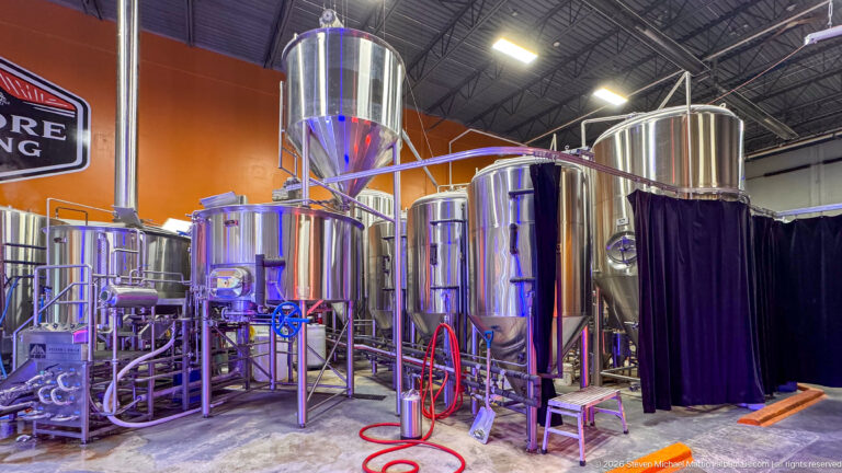 Stainless brewing tanks, a tall hopper, pipes, valves, and hoses occupy a high-ceilinged production room.