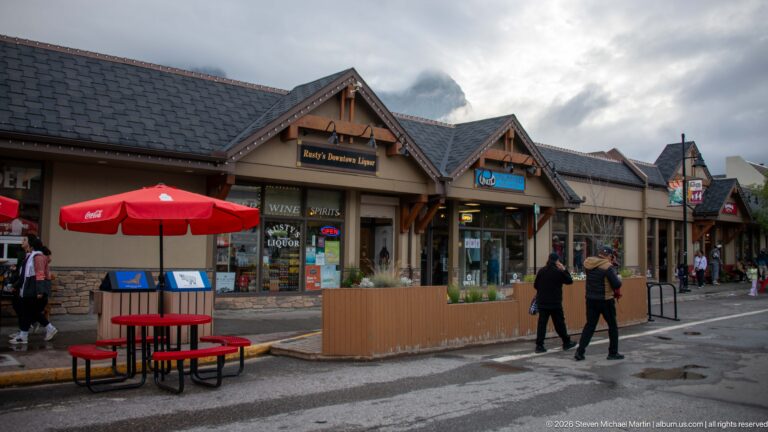 Timber-gabled shops and an outdoor seating area line a Canmore sidewalk below clouded peaks.