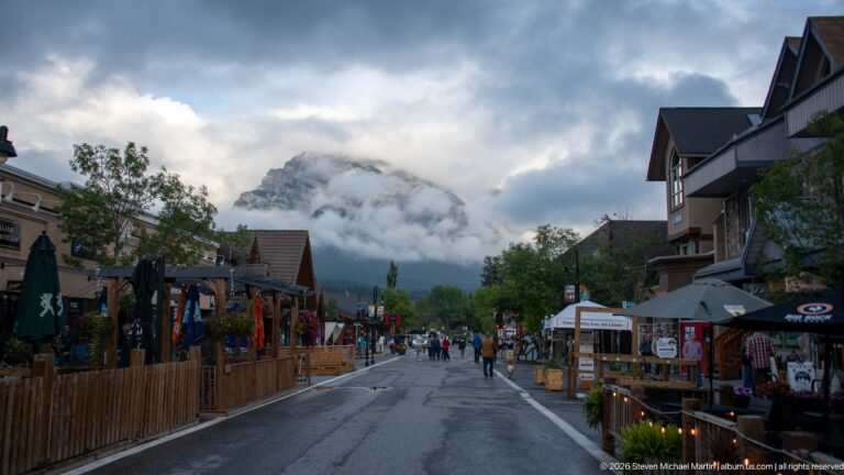 Visitors walk between timber-fronted shops along a pedestrian street beneath cloud-covered mountains.