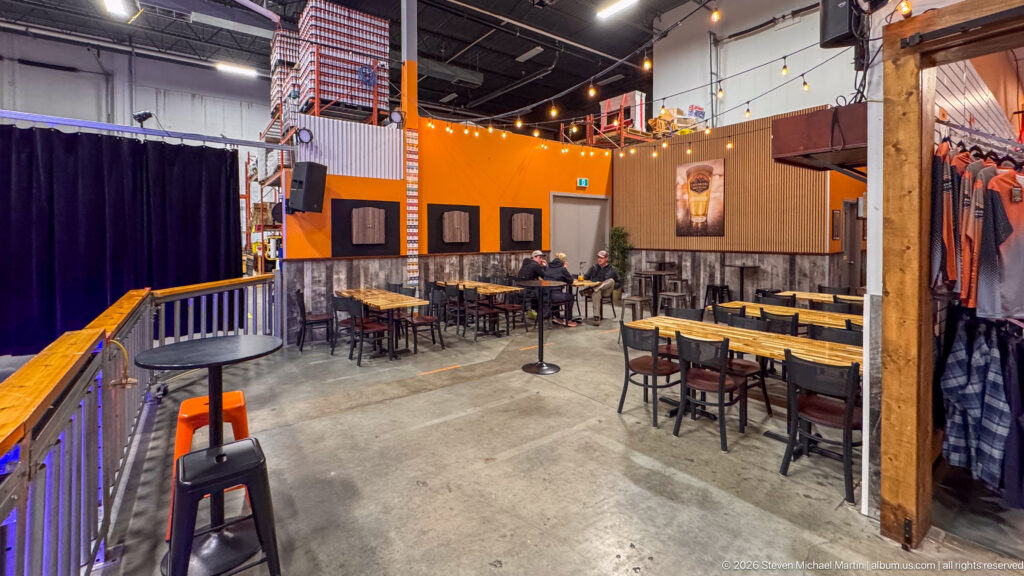Wooden tables and black stools fill an orange-walled brewery taproom, with seated patrons and hanging shirts.