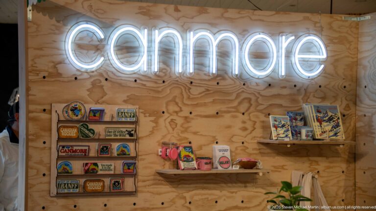 A white neon Canmore sign hangs above shelves of souvenirs on a plywood wall.