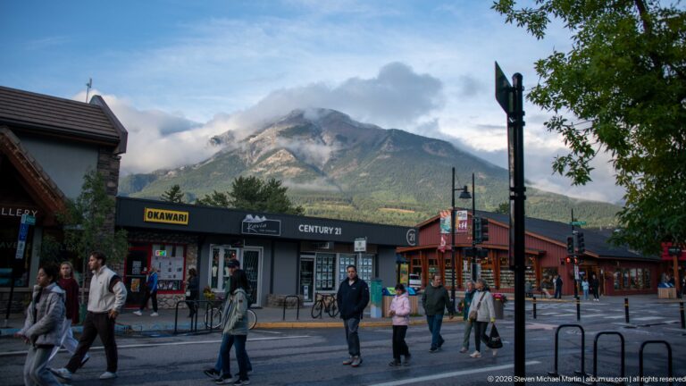 Pedestrians cross a commercial street beneath a clouded mountain, with storefronts along the block.