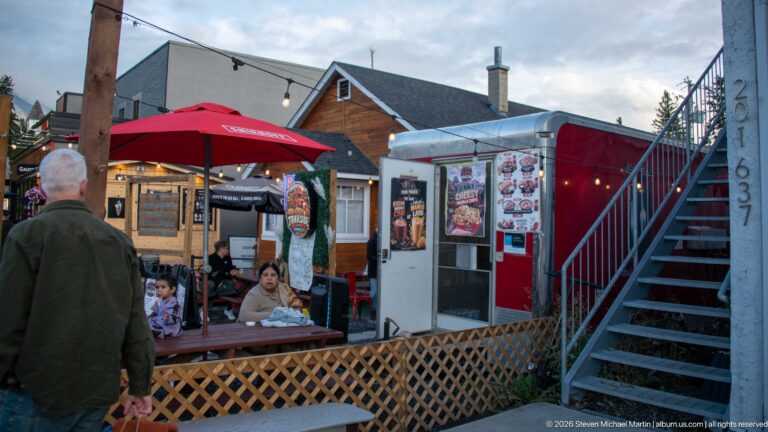A red food trailer serves an outdoor dining area with picnic tables and a red umbrella.