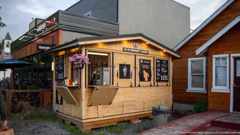 A timber food kiosk labeled Al’s Chimney Cakes stands along a narrow commercial lane.