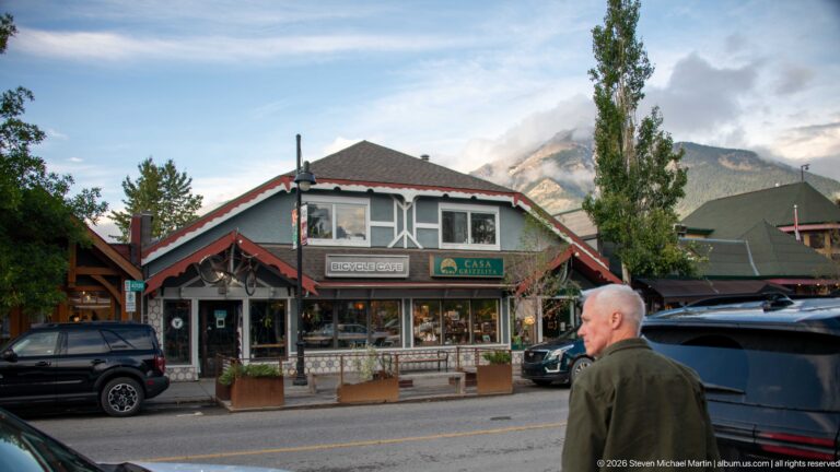 A pedestrian passes storefronts, including a Café Books sign, beneath a mountain ridge.