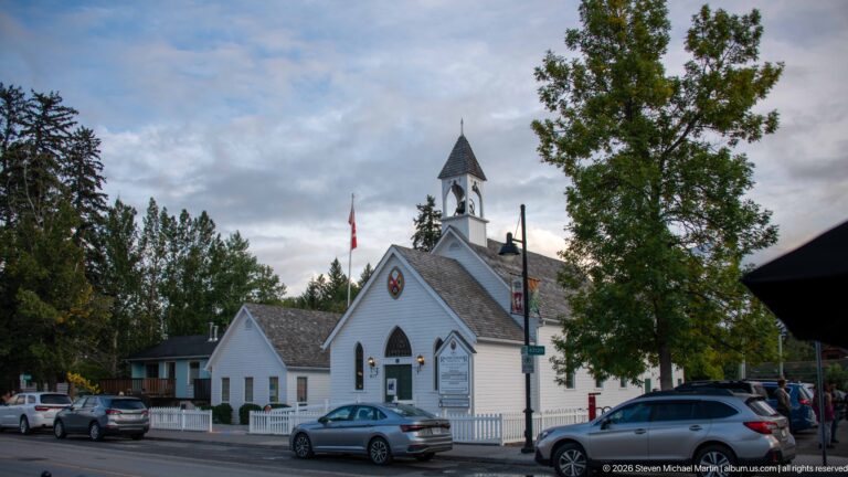 A white church and bell tower stand behind trees, a picket fence, and parked cars.