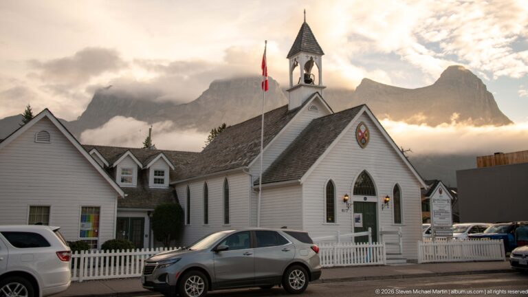 A white timber church with pointed windows and an open bell tower stands before the mountains.