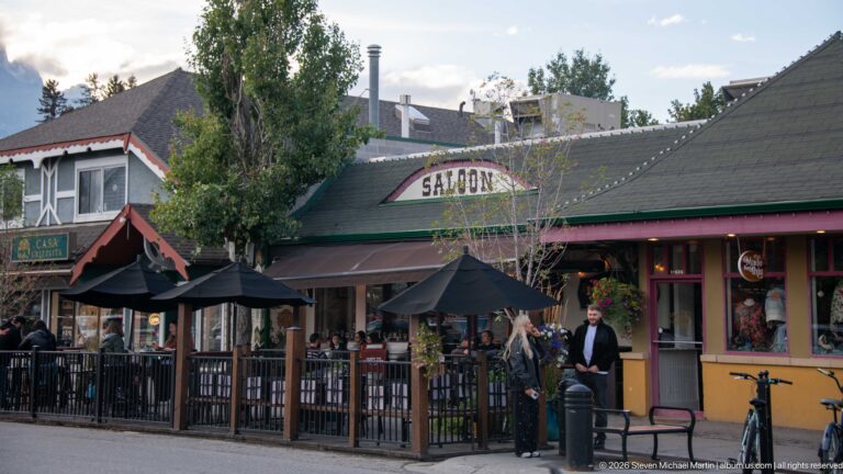 A saloon storefront opens onto a sidewalk patio with umbrellas, flower boxes, and pedestrians.