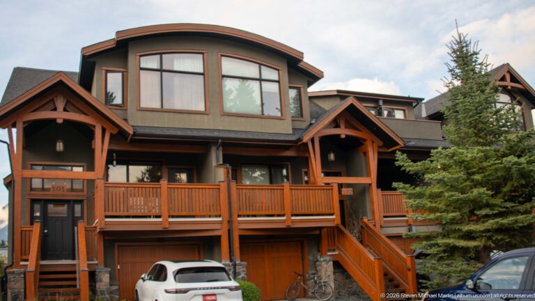 A townhouse with timber balconies and a street-level garage stands beside evergreen trees.