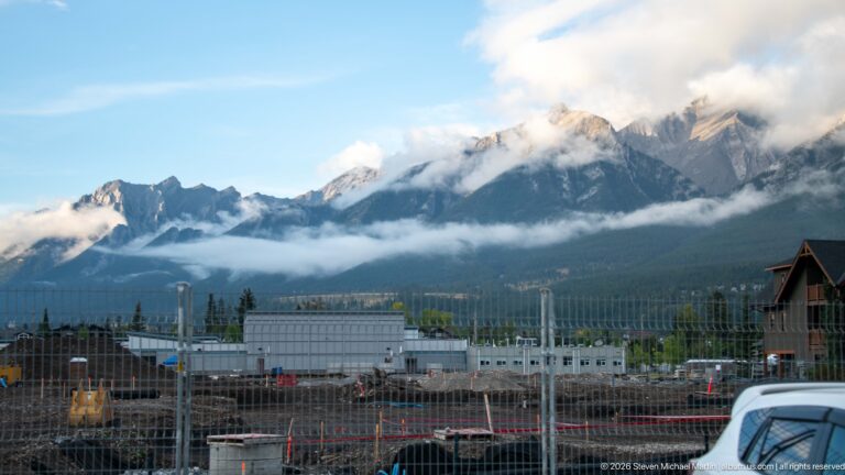 A fenced construction site and parked vehicles sit below a broad mountain slope wrapped in cloud.