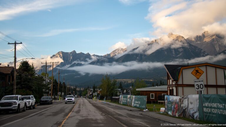 A residential road leads toward jagged, cloud-wrapped peaks beyond parked vehicles and construction fencing.